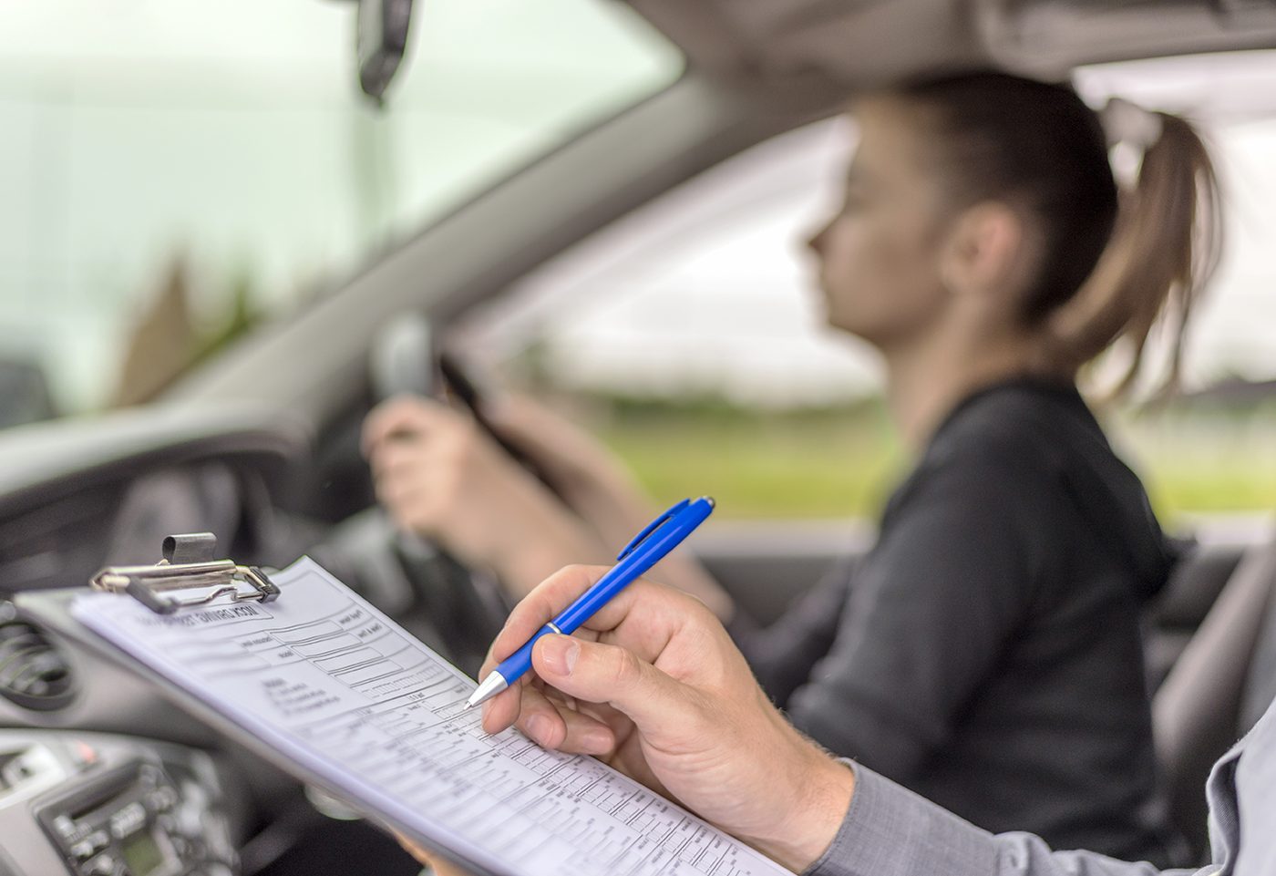 Driving instructor marking a test sheet while a learner drives a right-hand-drive car